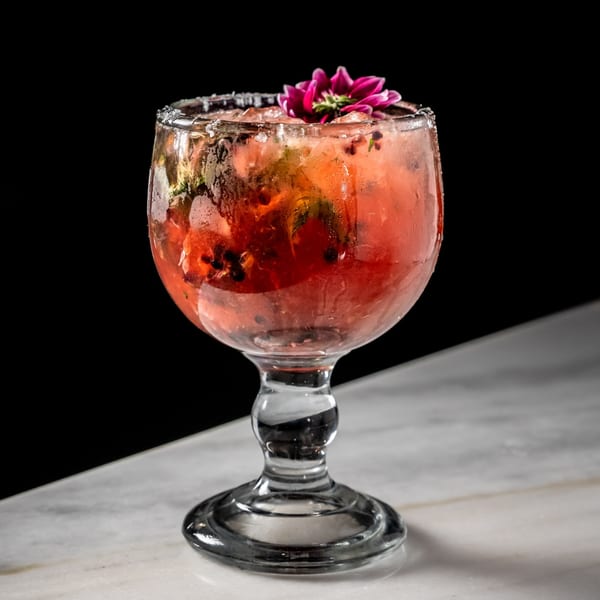 A vibrant red cocktail with ice, mint, and a pink flower garnish in a stemmed glass.