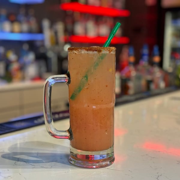 A large glass mug of a michelada with a chili-salted rim and a green straw on a bar.