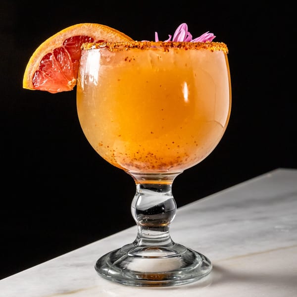 Orange cocktail in a goblet, garnished with grapefruit, chili-lime rim, and purple flowers.