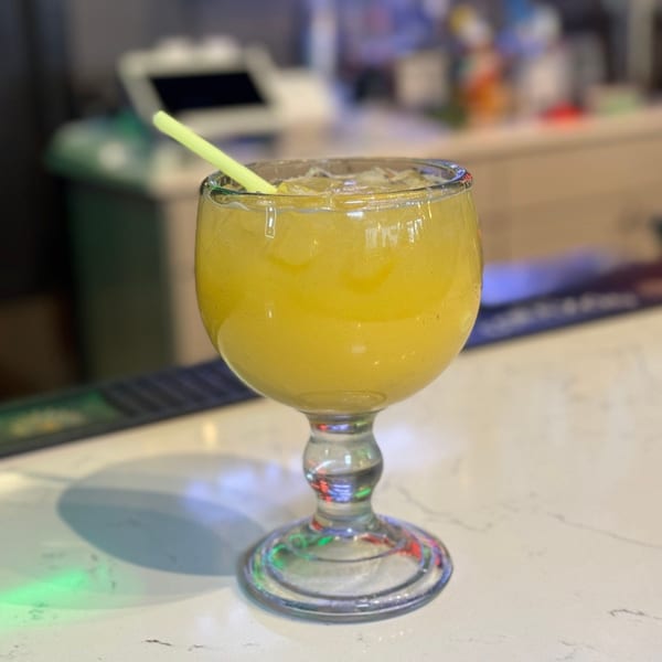 A large goblet glass filled with a yellow frozen drink and a green straw on a white bar.