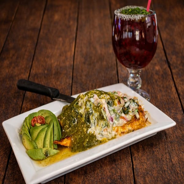 Grilled chicken with green and white sauce, avocado, and a red drink on a wooden table.