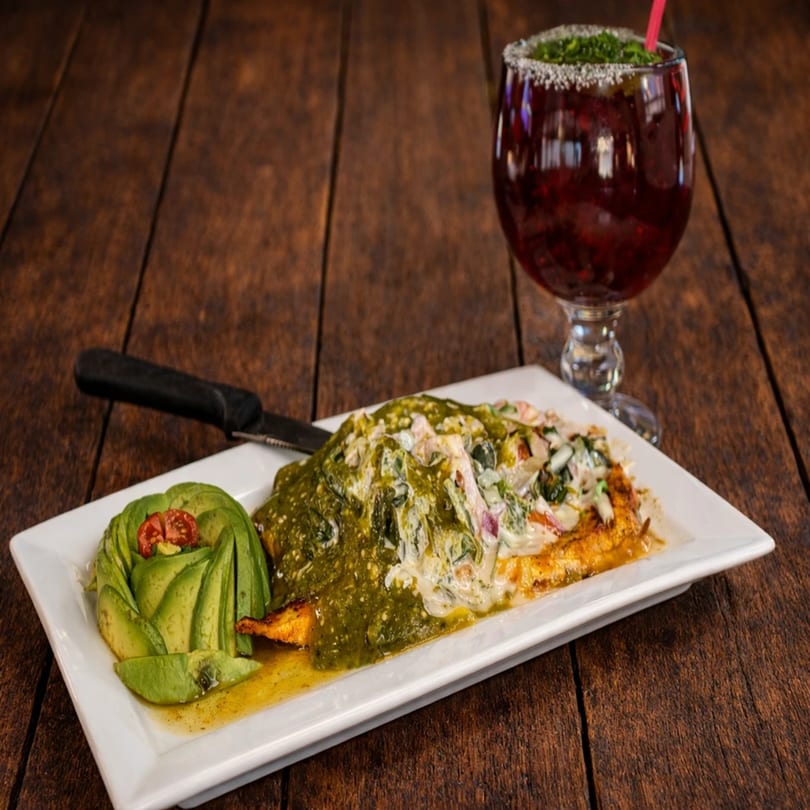 Grilled chicken with green and white sauce, avocado, and a red drink on a wooden table.