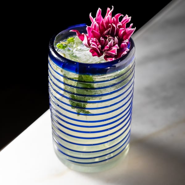 A vibrant cocktail in a blue and white striped glass, garnished with a large pink flower.