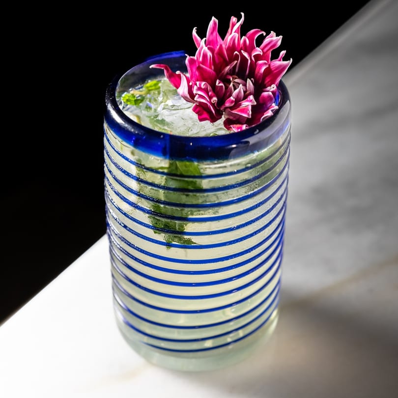 A vibrant cocktail in a blue and white striped glass, garnished with a large pink flower.